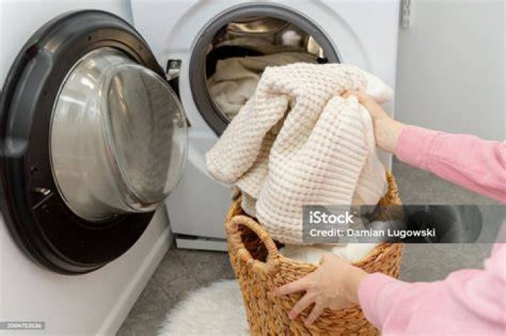 Woman putting white sweater clothes into the drum of a washing machine, front view.